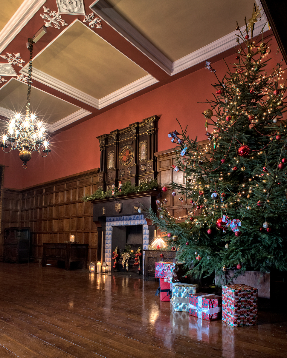Elegant room with wooden paneling and a high decorated ceiling, featuring a large Christmas tree with lights and ornaments, a grand chandelier, a garland-adorned fireplace, and wrapped presents beneath the tree.